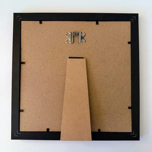 May include: The back of a black picture frame with a brown backing. A metal hanger is at the top, and a brown easel stand is attached to the back. The frame is square and has small clips to hold the backing in place.