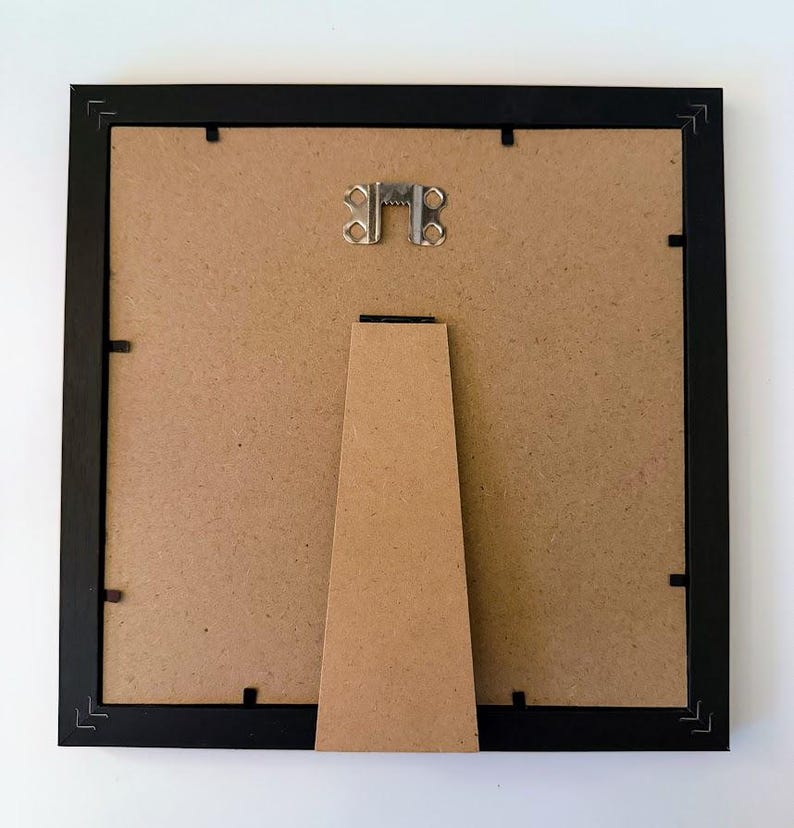 May include: The back of a black picture frame with a brown backing. A metal hanger is at the top, and a brown easel stand is attached to the back. The frame is square and has small clips to hold the backing in place.