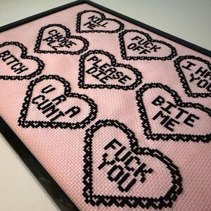 Puede incluir: Obra de arte de punto de cruz enmarcada con corazones negros sobre un fondo rosa. Los corazones muestran frases como "Bitch", "U R A Cunt" y "Fuck You". La obra est&aacute; enmarcada en negro.