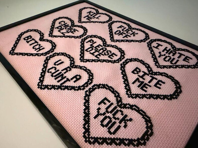 Puede incluir: Obra de arte de punto de cruz enmarcada con corazones negros sobre un fondo rosa. Los corazones muestran frases como "Bitch", "U R A Cunt" y "Fuck You". La obra est&aacute; enmarcada en negro.