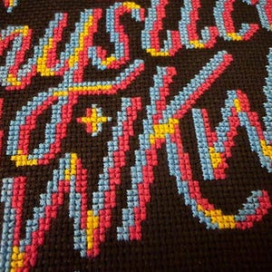 May include: Close-up of a cross-stitch textile with the word "JUSTICE" in colorful thread. The letters are primarily pink, blue, and yellow against a dark brown background. The stitching is detailed and the text is the focal point.
