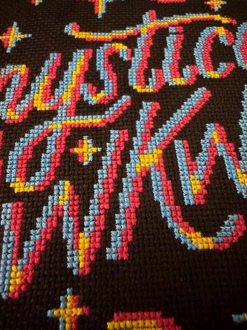 May include: Close-up of a cross-stitch textile with the word "JUSTICE" in colorful thread. The letters are primarily pink, blue, and yellow against a dark brown background. The stitching is detailed and the text is the focal point.