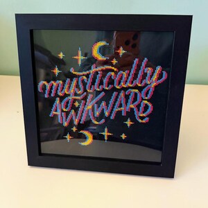 May include: A framed cross-stitch piece with the words "mystically awkward" in colorful thread. The text is in a stylized font with a 3D effect, surrounded by stars and crescent moons. The frame is black.