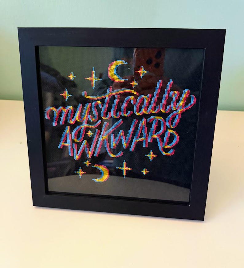 May include: A framed cross-stitch piece with the words "mystically awkward" in colorful thread. The text is in a stylized font with a 3D effect, surrounded by stars and crescent moons. The frame is black.