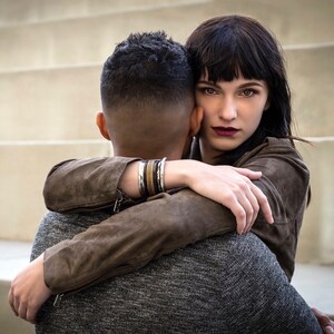 May include: A woman with dark hair and a brown leather jacket hugs a man from behind. She is looking at the camera with a serious expression. The man is wearing a gray sweater and has his back to the camera.