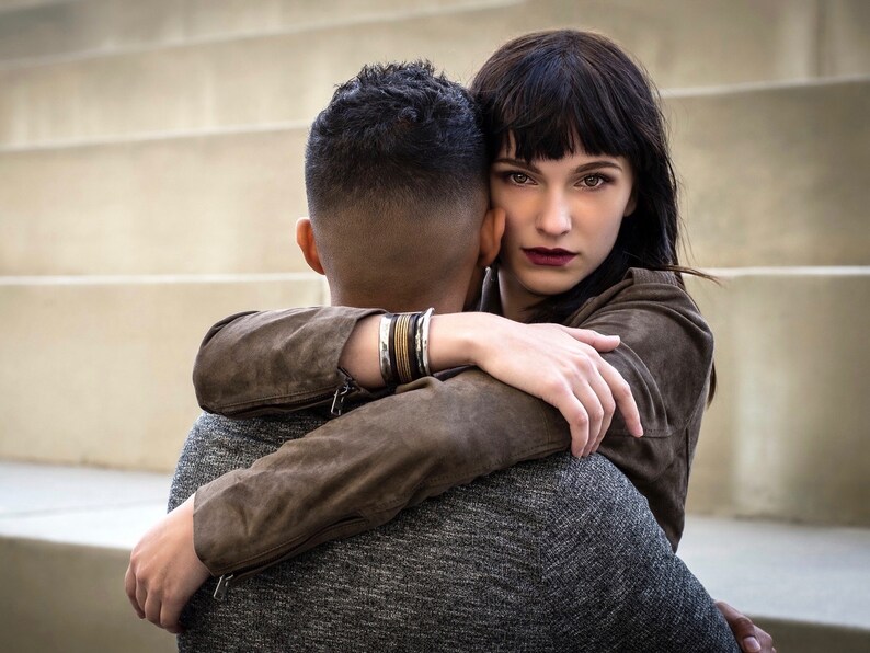 May include: A woman with dark hair and a brown leather jacket hugs a man from behind. She is looking at the camera with a serious expression. The man is wearing a gray sweater and has his back to the camera.