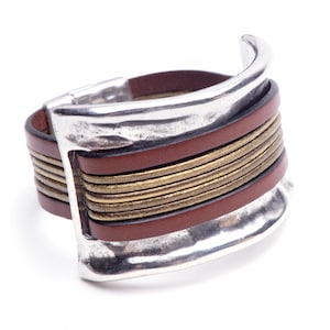 May include: A brown leather bracelet with a silver metal clasp and gold accents. The bracelet is wide and has a textured surface.