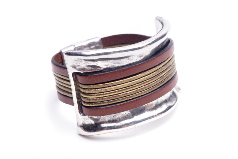 May include: A brown leather bracelet with a silver metal clasp and gold accents. The bracelet is wide and has a textured surface.
