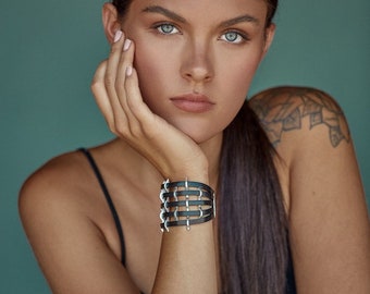 May include: A woman with long dark hair is wearing a black leather bracelet with silver accents. The bracelet is on her left wrist and has a geometric design.
