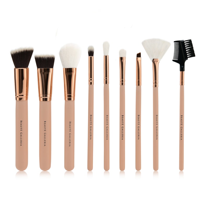 Op de afbeelding: Set van negen make-up kwasten met ros&eacute;gouden accenten en witte haren. De kwasten zijn in een rij op een witte achtergrond gerangschikt. De merknaam "BEAUTE GALLERIA" is op de handvatten van elke kwast gedrukt.
