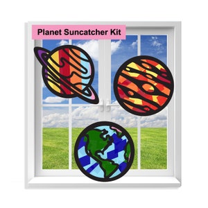 May include: A Planet Suncatcher Kit with three colorful planets: a blue and green Earth, a red and yellow Jupiter, and a rainbow-colored Saturn with rings. The planets are displayed against a window with a view of a green field and a blue sky.