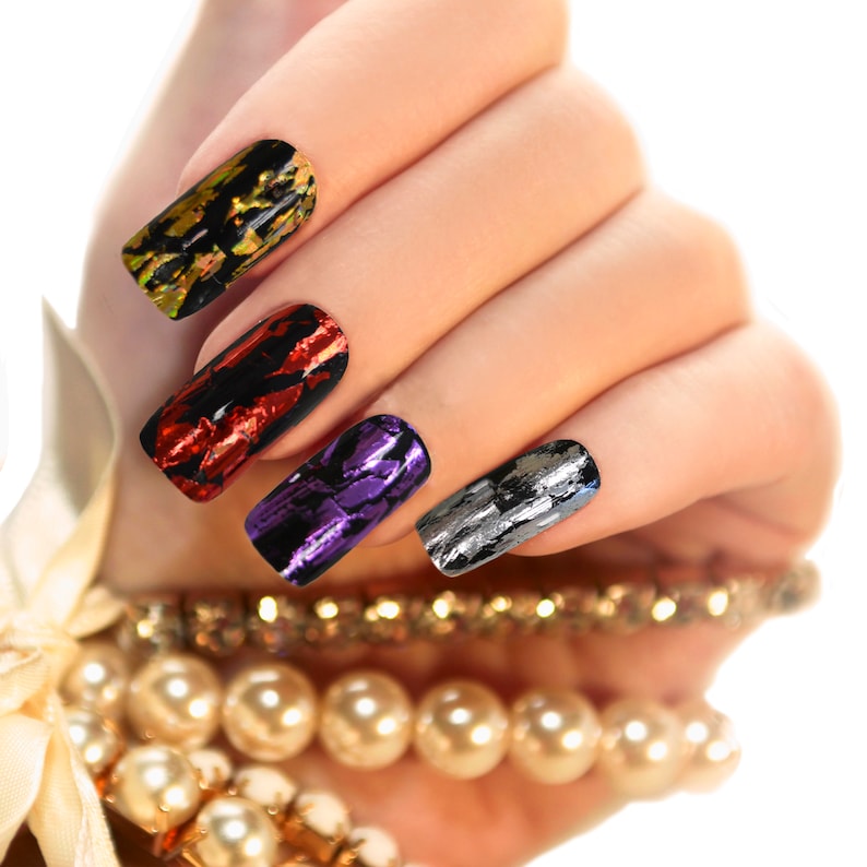 May include: Close-up of a hand with four fingernails painted with different colored nail polish. The nails are decorated with a foil-like finish in shades of gold, red, purple, and silver.