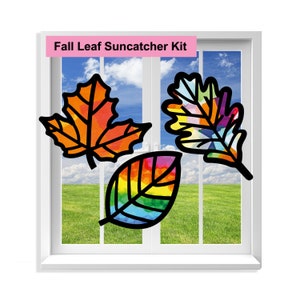 May include: A Fall Leaf Suncatcher Kit featuring three colorful leaf designs. The leaves are outlined in black and filled with vibrant hues of orange, yellow, and rainbow colors. The leaves are displayed in a window with a blue sky and green grass background.