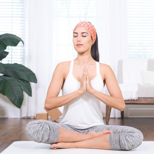 May include: A woman sits in a yoga pose with her eyes closed and hands in prayer position. She is wearing a white tank top, grey yoga pants, and a pink and orange headband.
