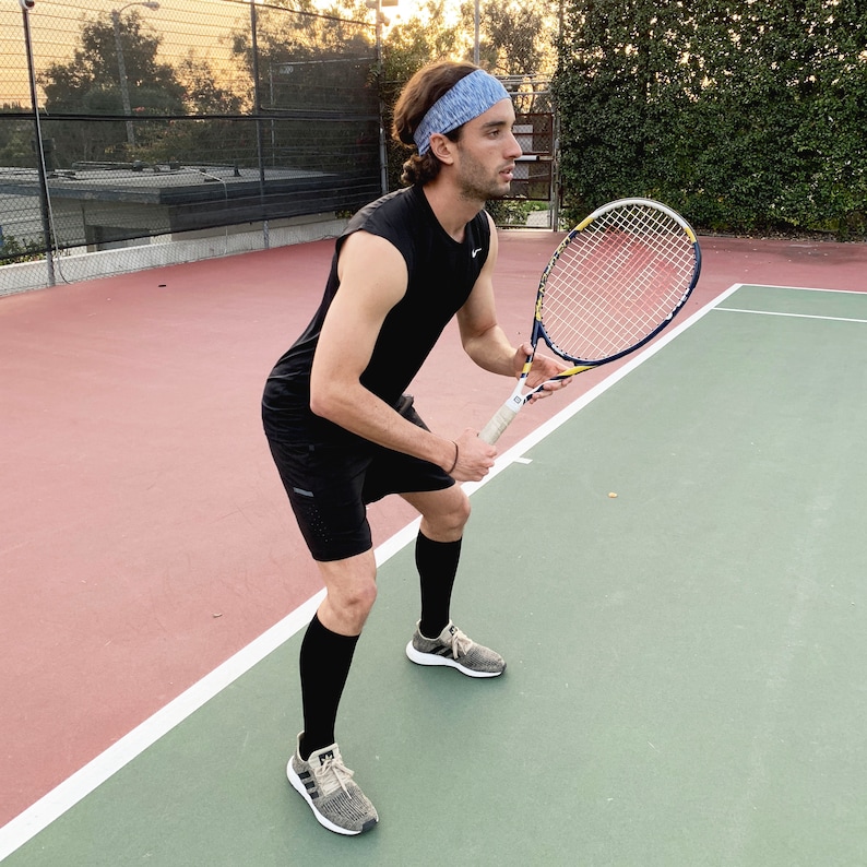 May include: A person wearing a black tank top and black shorts is holding a tennis racket on a tennis court. The person is wearing a blue headband and black socks with grey and white sneakers.