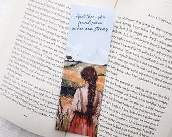 May include: Bookmark with a watercolor illustration of a woman with long brown hair looking at a field and a house. The bookmark has the text "And then, she found peace in her own storms" in a handwritten font. The bookmark is placed in an open book.