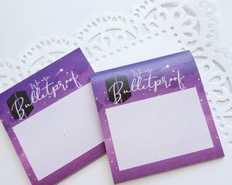May include: Two purple and white sticky note pads with the text "We are Bulletproof" in white lettering. The background of the note pads features a purple gradient with white clouds and gold stars.