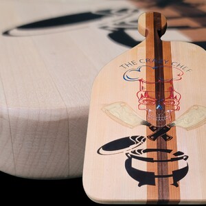 Puede incluir: Una tabla de cortar de madera con un diseño de calavera y huesos cruzados. La tabla está hecha de madera de arce y nogal y tiene un acabado natural. El texto "The Crazy Chef" está grabado en la tabla.