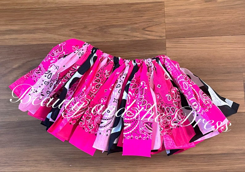 May include: A pink and black bandana skirt with a white paisley pattern. The skirt is made of multiple bandanas sewn together and has a gathered elastic waistband.