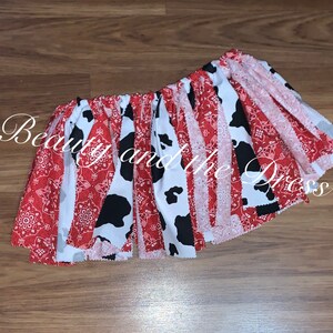 May include: A red and white cow print fabric skirt with red bandana trim. The skirt is gathered at the waist and has a ruffled hem.