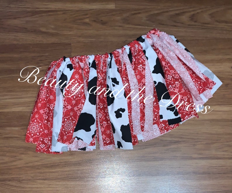 May include: A red and white cow print fabric skirt with red bandana trim. The skirt is gathered at the waist and has a ruffled hem.