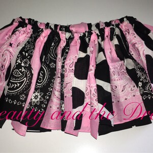 May include: A pink and black tutu skirt with a cow print and paisley pattern. The skirt is made of fabric scraps and has a gathered elastic waistband.