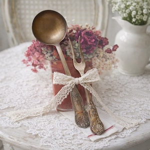 May include: A metal ladle and a two-pronged serving fork with ornate, embossed handles featuring shell-like motifs. The two utensils are tied together with a cream-coloured lace ribbon, creating a decorative set with a tarnished, aged finish.
