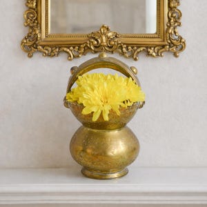 May include: A brass vessel with a rounded base, a narrow neck, and a wide top opening featuring a curved handle. The metal surface displays an etched pattern and a weathered patina. Inside the vessel sits a cluster of bright yellow flowers.