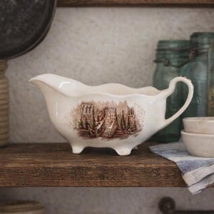 May include: A cream-coloured ceramic gravy boat featuring a brown transferware print of a building with a staircase. The vessel has a scalloped rim, a curved handle, and stands on three small feet, showcasing a detailed architectural scene.