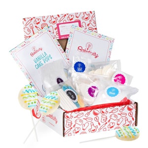 May include: A white box with a red and white pattern featuring a variety of baking tools. The box contains a baking kit for making vanilla cake pops. The kit includes a bag of cake mix, a bag of frosting, a bag of sprinkles, a bag of lollipop sticks, and a bag of candy melts. The box also includes two decorated cake pops on sticks.