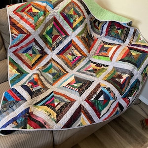 May include: A patchwork quilt featuring a repeating square block pattern composed of multi-colored fabric strips in various textures and prints. The blocks are separated by light gray grid-patterned sashing, and the quilt has a solid light green backing.