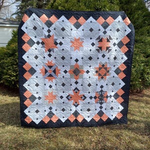 May include: A quilt with a black, white, and peach color scheme. The quilt is made up of a variety of geometric shapes, including squares, diamonds, and stars. The quilt is hanging on a clothesline in a grassy area.