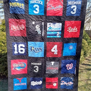 May include: A black and white quilt made from baseball jerseys. The quilt is made up of 16 squares, each with a different jersey. The jerseys are from different teams, including the Padres, Rays, Reds, and D'backs. The quilt is hanging on a clothesline in a backyard.