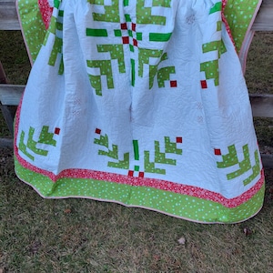 May include: A white quilt with a green and red Christmas tree design. The quilt has a red border with white snowflakes and a green border with white stars. The quilt is draped over a wooden fence.