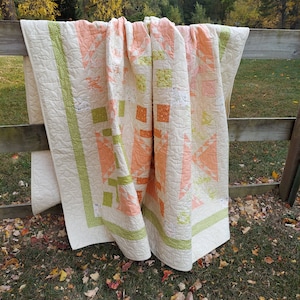 May include: A white quilt with a green and peach patchwork design. The quilt is hanging on a wooden fence with a green grassy background.