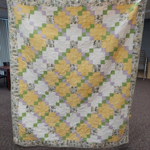 Pode incluir: Um edredom feito à mão com um design de patchwork. O edredom apresenta um padrão de quadrados amarelos, brancos e lavanda, acentuados com quadrados verdes. A borda é uma estampa floral. O edredom é exibido pendurado.