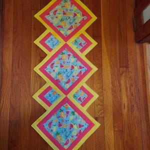 Peut inclure: Chemin de table matelassé avec un motif de losanges répétés. Chaque losange présente un fond bleu avec des motifs de pastèque et d'ananas. Le chemin de table a une bordure rose et un bord extérieur jaune. Le texte "Life is Sweet" est imprimé sur le tissu.