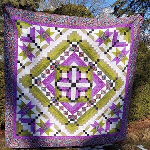 May include: A quilt with a geometric pattern in shades of green, purple, and white. The quilt is made up of squares and triangles, and the centre of the quilt features a large cross.