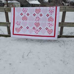 May include: A white quilt with a red border and a pattern of pink hearts with different designs. The quilt is hanging on a wooden fence against a snowy background.