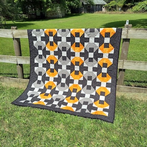 May include: A handmade quilt with a geometric pattern in black, gray, white, and orange. The quilt is draped over a wooden fence in a grassy outdoor setting. The quilt's design features repeating shapes.