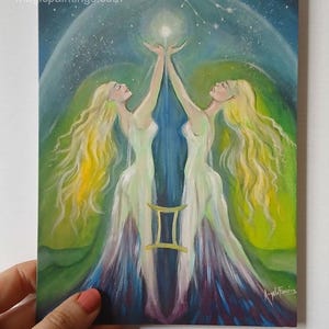 May include: A painting depicting two figures with long blonde hair and white gowns, reaching towards a bright light. The artwork includes a Gemini symbol and a starry background, with the website address magicpaintings.com visible.