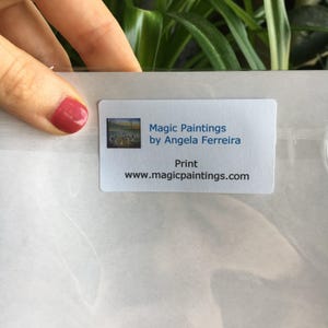 Puede incluir: Una pegatina rectangular blanca con el texto "Magic Paintings by Angela Ferreira" y "Print www.magicpaintings.com". La pegatina tiene una peque&ntilde;a imagen de una pintura. Una mano con esmalte de u&ntilde;as rojo sostiene la pegatina.
