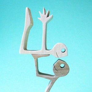 May include: A light-colored wooden sculpture depicting two stylized figures. One figure is supporting another figure above it. The upper figure has a hand-like shape. The sculpture is set against a light blue background.
