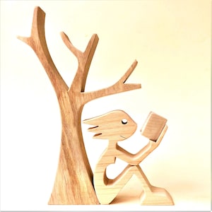 Könnte beinhalten: Holzskulptur einer Person, die unter einem Baum sitzt und ein Buch liest. Die Skulptur ist aus hellem braunem Holz gefertigt und hat ein schlichtes, stilisiertes Design.