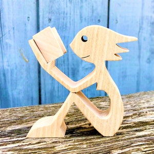 Peut inclure: Figurines en bois d'une personne lisant un livre. La figurine est en bois brun clair et a un style fantaisiste et caricatural. La personne est assise les jambes croisées et tient un livre dans ses mains.
