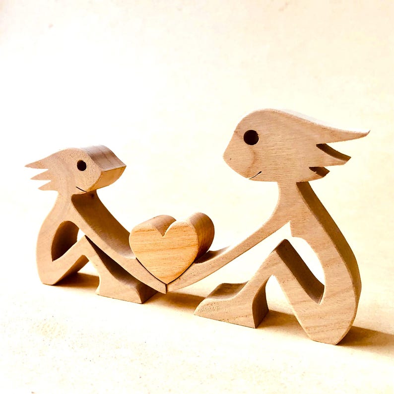K&ouml;nnte beinhalten: Holzskulptur von zwei Figuren, die ein Herz halten. Die Figuren sind in einem einfachen Stil geschnitzt und haben eine hellbraune Farbe.