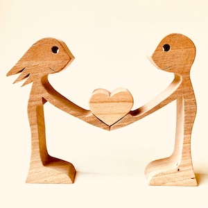May include: Wooden sculpture of two figures holding a heart. The figures are stylized with simple shapes and lines, and the heart is made of the same light-colored wood. The sculpture is a symbol of love and connection.