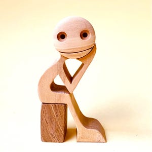 Può includere: Una scultura in legno di una persona in posizione seduta, con una mano che sostiene il mento. La figura ha una testa rotonda con due fori per gli occhi e un sorriso scolpito. La scultura è realizzata in legno marrone chiaro e poggia su un piccolo blocco.