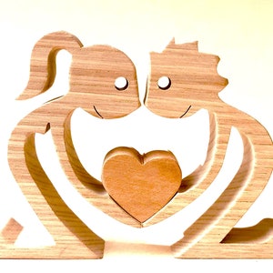May include: Wooden sculpture of two figures holding a heart in the center. The figures are carved in a simple style with rounded features and are made of light brown wood.