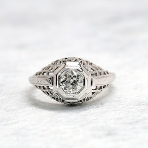 May include: A vintage white gold engagement ring with a round brilliant cut diamond set in a delicate filigree design.